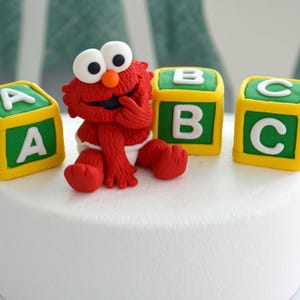 May include: A red Elmo character figurine made of fondant sitting on a white cake. The figure has large white eyes with black pupils and an orange nose. It is surrounded by three green and yellow square blocks featuring the white letters A, B, and C.