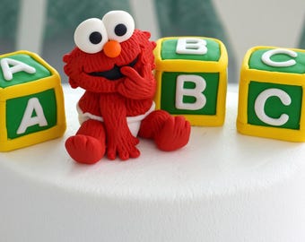 May include: A red Elmo character figurine made of fondant sitting on a white cake. The figure has large white eyes with black pupils and an orange nose. It is surrounded by three green and yellow square blocks featuring the white letters A, B, and C.