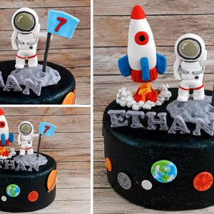以下が含まれることがあります： 宇宙をテーマにしたフォンダンフィギュアで飾られた黒いケーキ。白いロケット、数字の7が書かれた青い旗を持つ宇宙飛行士、ETHANという文字を形作る岩のような質感の灰色の文字が含まれています。側面には平らな惑星の装飾が施されています。