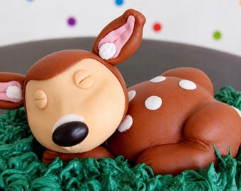 Op de afbeelding: Een bruin fondant hertje met een lichtbruin gezicht, gesloten ogen en een zwarte neus. Het hertje heeft roze gevoerde oren, witte stippen op de rug en rust op een bedje van groen gespoten glazuur dat op gras lijkt.