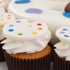 May include: A cupcake with a brown paper liner, topped with white frosting and a white, flat, circular artist palette decoration. The palette features small, colourful dots in red, orange, yellow, green, blue, and purple, mimicking paint colours.
