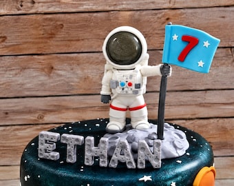 Op de afbeelding: Een ronde taart met een donkere ruimte-afwerking met een wit astronautfiguurtje dat op een grijs oppervlak met kraters staat. De astronaut houdt een lichtblauwe vlag vast met een rood cijfer 7 en witte sterren. De naam ETHAN staat in grijze letters.