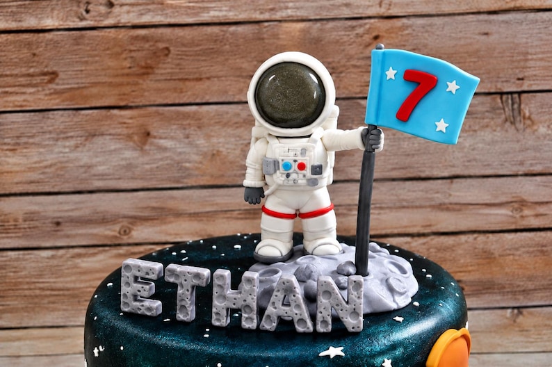 May include: A round cake with a dark space-themed finish featuring a white astronaut figurine standing on a gray cratered surface. The astronaut holds a light blue flag with a red number 7 and white stars. The name ETHAN is spelled out in gray letters.