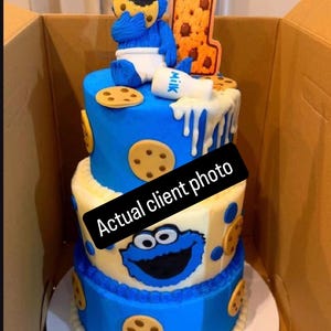 May include: A three-tier cake decorated with blue and white frosting, featuring a blue monster figure eating a cookie, a white milk bottle, and a large number one topper. The cake is adorned with cookie cutouts and a black sign reading Actual client photo.