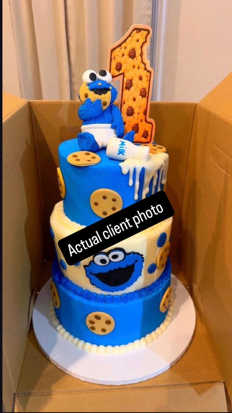 May include: A three-tier cake decorated with blue and white frosting, featuring a blue monster figure eating a cookie, a white milk bottle, and a large number one topper. The cake is adorned with cookie cutouts and a black sign reading Actual client photo.