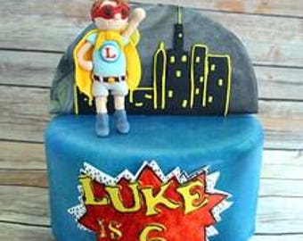 Op de afbeelding: Een ronde blauwe taart met een fondant superheldfiguurtje met een gele cape en een rood masker. De taart is bekroond met een zwart silhouet van een stad. De voorkant toont een rood-witte stripstijl explosie met de tekst LUKE IS 6.
