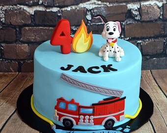 Op de afbeelding: Een lichtblauwe ronde taart versierd met een rood cijfer 4, een gele en oranje vlam en een wit puppyfiguurtje met zwarte stippen. De zijkant toont een rode brandweerwagen, een grijze ladder, de naam JACK en een blauwe Happy Birthday-banner.