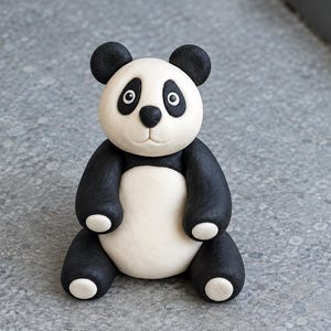 Może przedstawiać: Mała, siedząca figurka pandy o gładkim, matowym wykończeniu. Panda ma biały pyszczek z czarnymi plamami wokół oczu, czarny nos i czarne uszy. Jej tułów jest biały, z czarnymi rękami i nogami, zakończonymi białymi łapami.