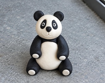 Op de afbeelding: Een klein, zittend panda-beeldje met een gladde, matte afwerking. De panda heeft een wit gezicht met zwarte vlekken rond de ogen, een zwarte neus en zwarte oren. Het lichaam is wit met zwarte armen en benen, elk eindigend in witte pootjes.