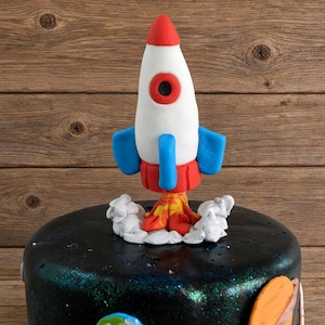 May include: A fondant rocket ship cake topper featuring a white body with a red nose cone, blue fins, and a red base. The rocket is surrounded by white fondant clouds and orange and yellow flames, positioned atop a black space-themed cake.