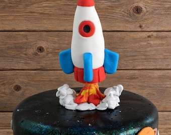 May include: A fondant rocket ship cake topper featuring a white body with a red nose cone, blue fins, and a red base. The rocket is surrounded by white fondant clouds and orange and yellow flames, positioned atop a black space-themed cake.