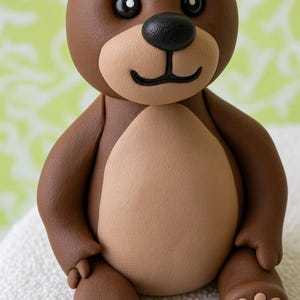 May include: A brown teddy bear figurine with a smooth matte finish. The bear features a lighter tan belly, muzzle, and paw pads. It has round ears, black bead-like eyes, and a black nose with a small curved mouth line. The bear is in a seated position.