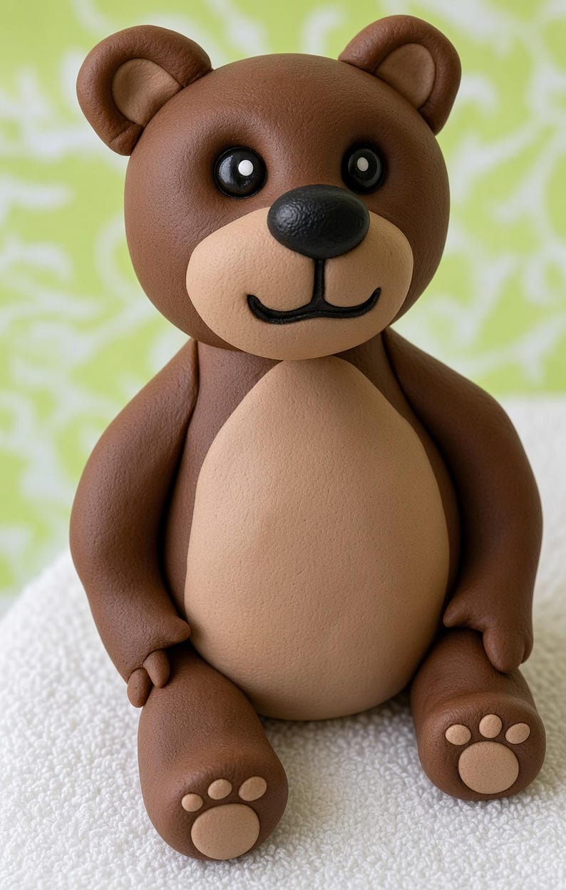 May include: A brown teddy bear figurine with a smooth matte finish. The bear features a lighter tan belly, muzzle, and paw pads. It has round ears, black bead-like eyes, and a black nose with a small curved mouth line. The bear is in a seated position.