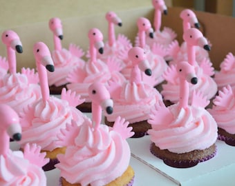Op de afbeelding: Een verzameling cupcakes in een doos, elk voorzien van een toef lichtroze glazuur en een klein, gevormd roze flamingo-figuurtje met een lange nek, een snavel met een zwarte punt en kleine roze vleugels aan de zijkanten.