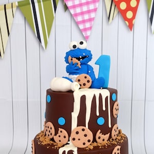 May include: A two-tier chocolate cake decorated with blue polka dots and chocolate chip cookie cutouts. A blue monster figurine sits on top eating a cookie next to a white milk pitcher and a blue number one. White icing drips down the sides.