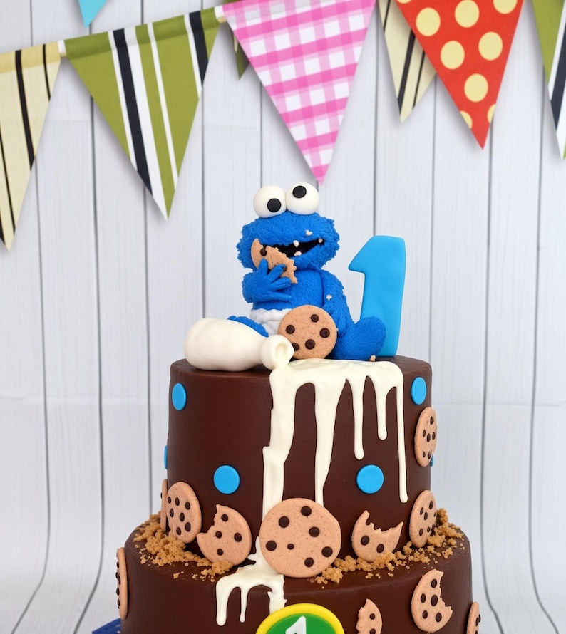 May include: A two-tier chocolate cake decorated with blue polka dots and chocolate chip cookie cutouts. A blue monster figurine sits on top eating a cookie next to a white milk pitcher and a blue number one. White icing drips down the sides.
