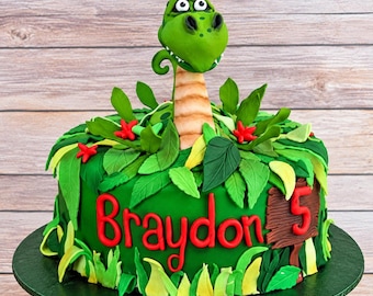 Op de afbeelding: Een ronde groene taart versierd met eetbare junglebladeren en rode bloemen. Een gebeeldhouwde groene dinosaurus kop en nek komen uit het midden. De naam Braydon staat in rode letters op de zijkant, naast een bruin bordje met het cijfer 5.