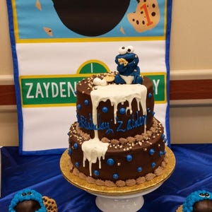 May include: A two-tier chocolate cake with white icing drips, blue dots, and brown frosting borders. A blue monster figurine eating a cookie sits on top. The cake is inscribed with "Birthday Zayden" and accompanied by two smaller blue frosted treats.