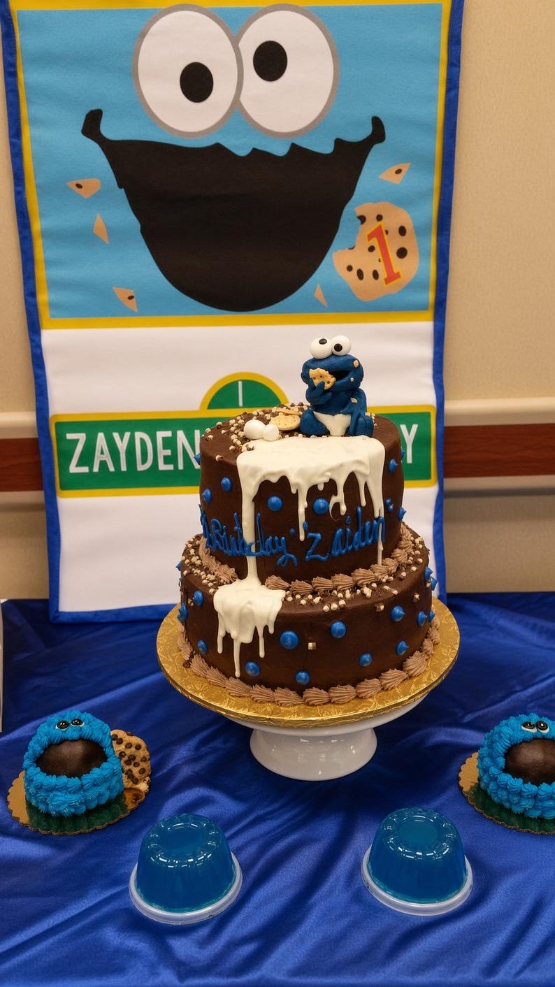 May include: A two-tier chocolate cake with white icing drips, blue dots, and brown frosting borders. A blue monster figurine eating a cookie sits on top. The cake is inscribed with "Birthday Zayden" and accompanied by two smaller blue frosted treats.