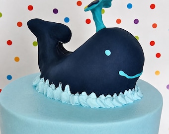 Op de afbeelding: Een donkerblauwe walvis taarttopper gemaakt van fondant, met een lichtblauwe waterstraal, een klein lichtblauw oog en een gebogen lichtblauwe mond. De walvis staat op een lichtblauwe taartbasis met lichtblauwe, gespoten glazuurgolven.