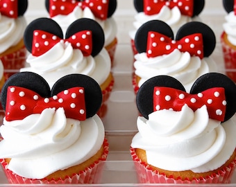 May include: A collection of cupcakes featuring white frosting swirls topped with black circular ears and red bows with white polka dots. Each cupcake sits in a red paper liner with a white polka dot pattern, arranged in a clear plastic tray.