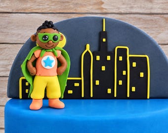 Op de afbeelding: Een set taartdecoraties van fondant met een superheldfiguur met een groen masker en cape, een oranje shirt met een blauwe ster en een gele broek. Achter de figuur staat een donkergrijze halfronde achtergrond met een zwart silhouet van een stad.