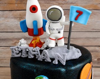 Op de afbeelding: Een taarttopper-set met ruimtethema bestaande uit een wit-rode raket met blauwe vinnen, een wit astronautfiguurtje en een lichtblauwe vlag met het rode cijfer 7. De set bevat grijze letters die de naam ETHAN vormen en kleine planeetdecoraties.