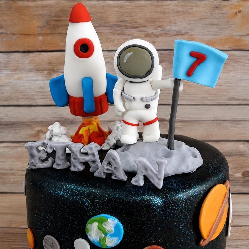 以下が含まれることがあります： 宇宙をテーマにしたケーキトッパーセット。青いフィンが付いた白と赤のロケット、白い宇宙飛行士のフィギュア、赤い数字の7が描かれた水色の旗が含まれています。グレーの文字でETHANと綴られたパーツと、小さな惑星の装飾が施されています。