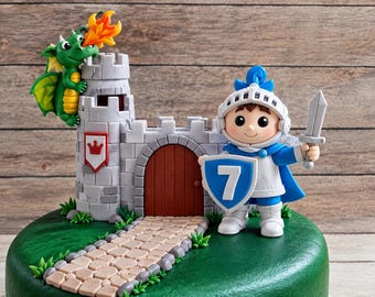 May include: A cake topper set featuring a gray stone castle with a red door, a green dragon breathing orange fire, and a knight in blue and white armor holding a gray sword and a blue shield with the white number 7, all on a green base.