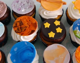 Op de afbeelding: Een verzameling cupcakes versierd met eetbare toppings met ruimtethema, waaronder planeten met gemarmerde texturen, een gele zon, een planeet met ringen en een zwarte schijf met gele sterren, allemaal op toeven van witte glazuur.