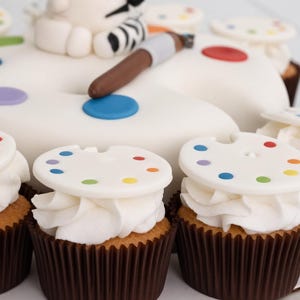 May include: A large white cake shaped like an artist palette with a brown paintbrush on top, surrounded by several cupcakes. Each cupcake is topped with white frosting and a small white disc decorated with tiny colourful paint spots.