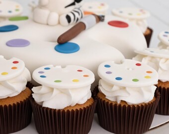 Op de afbeelding: Een grote witte taart in de vorm van een kunstenaarspalet met een bruine kwast erop, omringd door verschillende cupcakes. Elke cupcake is bedekt met witte glazuur en een kleine witte schijf versierd met kleine kleurrijke verfstippen.