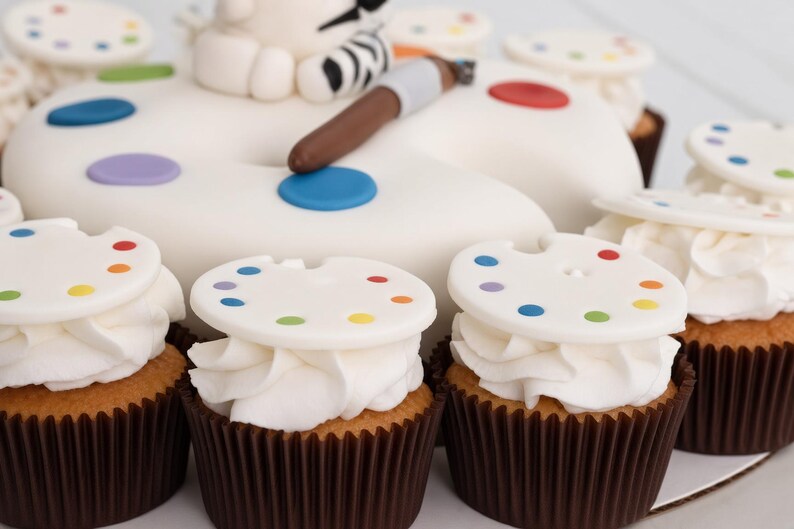 May include: A large white cake shaped like an artist palette with a brown paintbrush on top, surrounded by several cupcakes. Each cupcake is topped with white frosting and a small white disc decorated with tiny colourful paint spots.