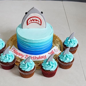 May include: A round cake with blue ombre frosting topped with a gray shark fin decoration featuring a red mouth and white teeth. A white banner with the red text Happy Birthday sits at the base, surrounded by six cupcakes with blue frosting.