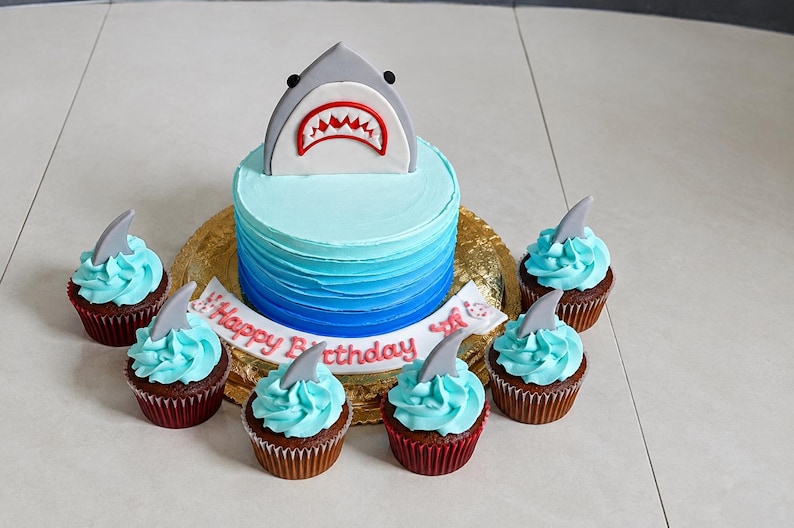 May include: A round cake with blue ombre frosting topped with a gray shark fin decoration featuring a red mouth and white teeth. A white banner with the red text Happy Birthday sits at the base, surrounded by six cupcakes with blue frosting.