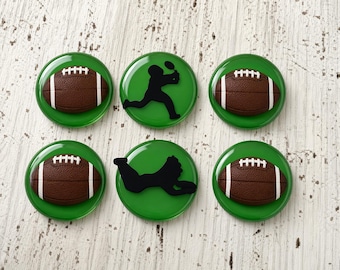 Op de afbeelding: Een set van zes ronde groene knopen met reliëf. Vier knopen tonen een bruine American football met witte veters, terwijl twee knopen een zwart silhouet van een footballspeler in verschillende sportieve houdingen bevatten.