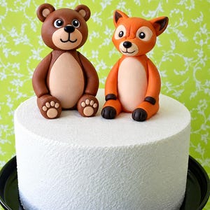 May include: A white cylindrical cake topped with two fondant animal figurines. On the left, a brown bear with a light tan belly and paw pads. On the right, an orange fox with a light tan belly, dark brown ear tips, and dark brown paws.