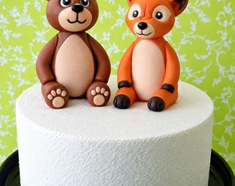 Op de afbeelding: Een witte cilindervormige taart met twee fondant dierenfiguurtjes bovenop. Links een bruine beer met een lichtbruine buik en voetzooltjes. Rechts een oranje vos met een lichtbruine buik en donkerbruine oortoppen en pootjes.