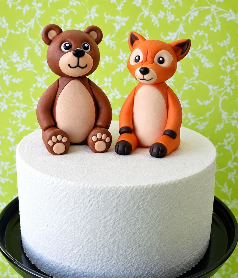 May include: A white cylindrical cake topped with two fondant animal figurines. On the left, a brown bear with a light tan belly and paw pads. On the right, an orange fox with a light tan belly, dark brown ear tips, and dark brown paws.