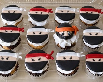 Op de afbeelding: Een set van twaalf cupcakes in een doorzichtige plastic verpakking. Elf cupcakes hebben fondant ninja-gezichten met zwarte maskers en rode of witte hoofdbanden. Eén centrale cupcake heeft een ninja-gezicht met een oranje hoofdband en ogen.