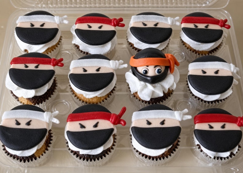 May include: A set of twelve cupcakes arranged in a clear plastic container. Eleven cupcakes feature fondant ninja faces with black masks and either red or white headbands. One central cupcake features a ninja face with an orange headband and visible eyes.