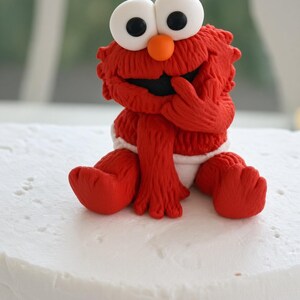 May include: A red, textured fondant figurine of a character with large white eyes, black pupils, and an orange nose. The figure is sitting down, wearing a white diaper, and has one hand raised to its mouth. The character has a fuzzy appearance.