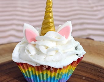 May include: A cupcake topped with white frosting, two small pink and white ears, and a central gold spiral horn. The cupcake is held in a pleated paper liner featuring horizontal stripes of red, orange, yellow, green, blue, and purple colors.