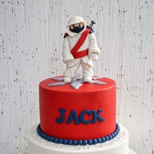 Może przedstawiać: Okrągły czerwony tort z białą figurką ninja z czerwoną szarfą i czarną maską na szczycie. Imię JACK jest napisane niebieskimi literami z przodu tortu. Niebieska krawędź z koralik&oacute;w otacza podstawę czerwonego poziomu, kt&oacute;ry stoi na białej podstawie.