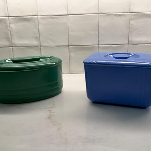 May include: A dark green oval ceramic butter dish with a matching lid and a bright blue rectangular ceramic butter dish with a matching lid. Both containers feature horizontal ribbed detailing along the sides and a smooth, glossy finish.
