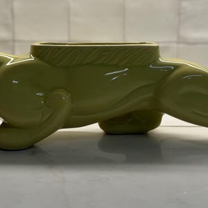 May include: A glossy, light olive green ceramic planter shaped like a prowling panther in a low, elongated stance. The sculpture features a smooth, reflective finish with a central rectangular opening on the back for holding small items or plants.