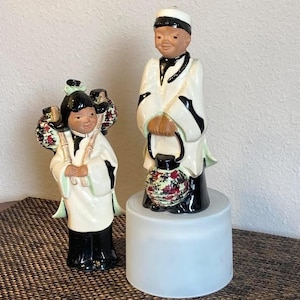 May include: Two ceramic figurines depicting individuals in traditional attire. One figure stands on a raised cylindrical base, holding a lantern with a floral pattern. The other figure stands beside it, carrying a floral-patterned basket on their back.