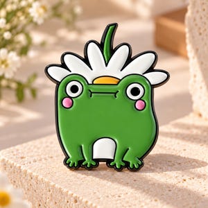 May include: A green frog enamel pin featuring a white daisy flower on its head. The frog has large black eyes, pink cheeks, and a white belly patch. The design is outlined in black metal, creating a clear, cartoon-style illustration of a frog.