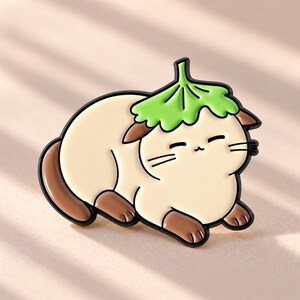 May include: A cream-colored enamel pin shaped like a cat lying down with brown ears, paws, and tail. The cat wears a bright green leaf-shaped hat on its head. The design features black outlines and a smiling expression with closed eyes and whiskers.