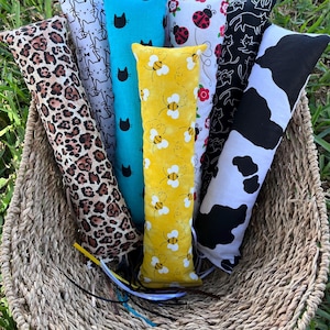 May include: A collection of six long, rectangular fabric catnip toys in a woven basket. The patterns include leopard print, white with black cats, solid turquoise with black cat heads, yellow with bees, white with ladybugs, and black and white cow print.